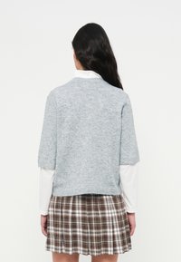 Ανοιχτό γκρι πλεκ τό πουλόβερ με κοντά μανίκια και στρογγυλή λαιμόκοψη, φορεμένο πάνω από λευκό μπλουζάκι με μακριά μανίκια, σε συνδυασμό με καφέ ριγέ πλισέ φούστα.