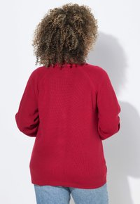 Roter Strickpullover mit strukturiertem Finish, ausgestattet mit Raglanärmeln und einem dekorativen Rüschendetail entlang des Ausschnitts.