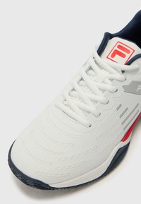 White athletic shoe with grey accents, textured surface, and red detailing. Features a lace-up closure and a navy tongue. Rubber sole for traction.