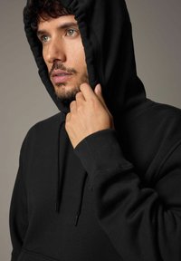 Schwarzer Kapuzenpullover mit Kordelzug, langen Ärmeln und gerippten Bündchen. Der Stoff wirkt weich und strukturiert und unterstreicht einen lässigen Look.