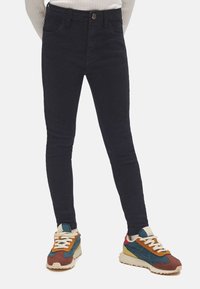 Vaqueros oscuros ajustados de cintura alta, con una textura suave. Combinados con zapatillas deportivas multicolores, con acentos en crema, azul y naranja.