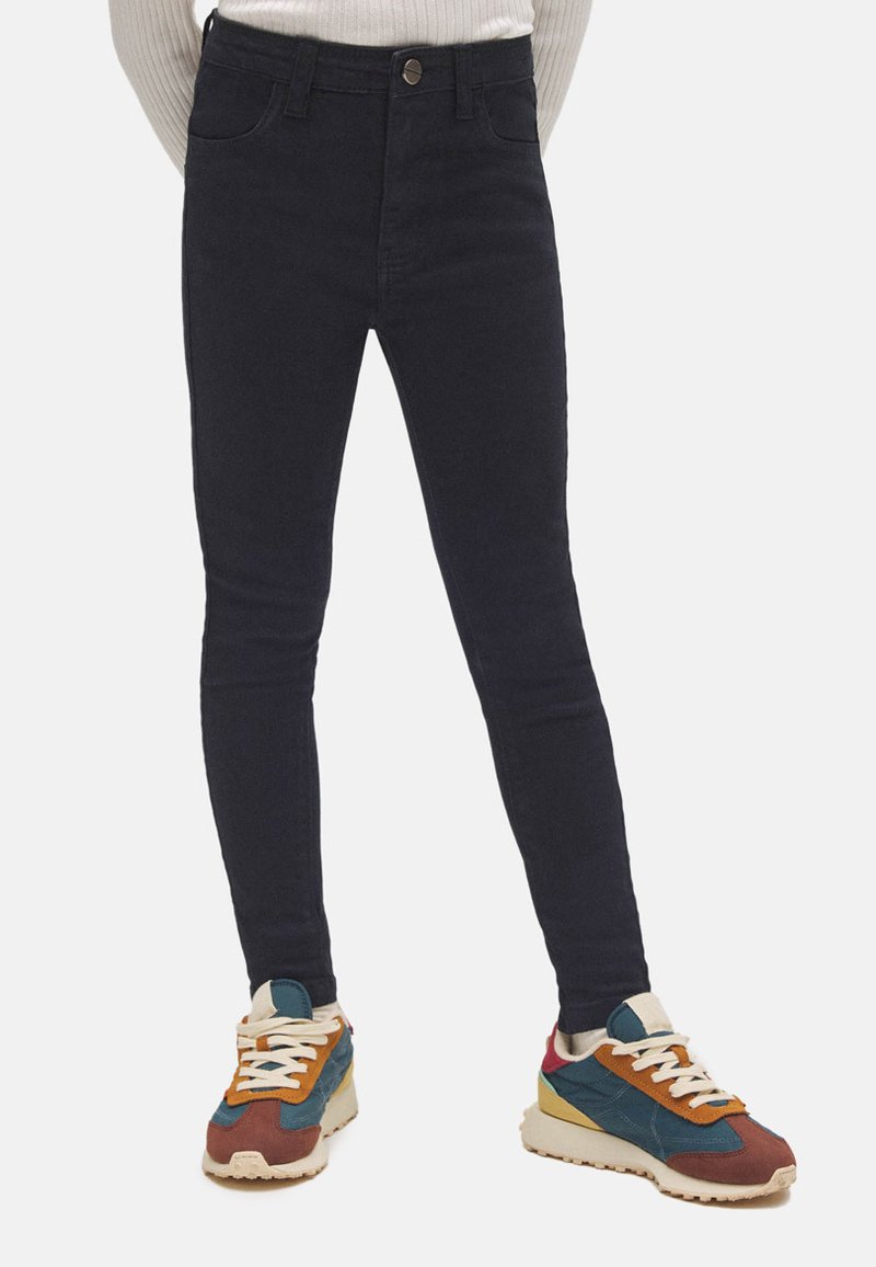 Vaqueros oscuros ajustados de cintura alta, con una textura suave. Combinados con zapatillas deportivas multicolores, con acentos en crema, azul y naranja.