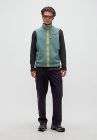 Νέος άνδρας με πράσινο γιλέκο φλις, μαύρο μακρυμάνικο πουκάμισο, μαύρο παντελόνι cargo, γυαλιά ηλίου και παπούτσια πεζοπορίας, στέκεται μπροστά σε απλό φόντο.