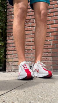 Baltas sporta apavus ar sarkaniem akcentiem, ar sietveida augšdaļu, biezu zoli un šņorēm. Novietots uz bruģētas virsmas pret ķieģeļu fona.