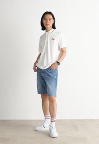 White polo shirt with red collar accents, denim shorts, white sneakers. Model has long hair, stands against a plain background.