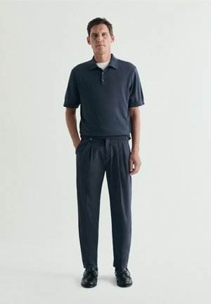 Hombre con camiseta polo azul marino, pantalones plisados a juego y zapatos negros, de pie frente a un fondo claro y liso.