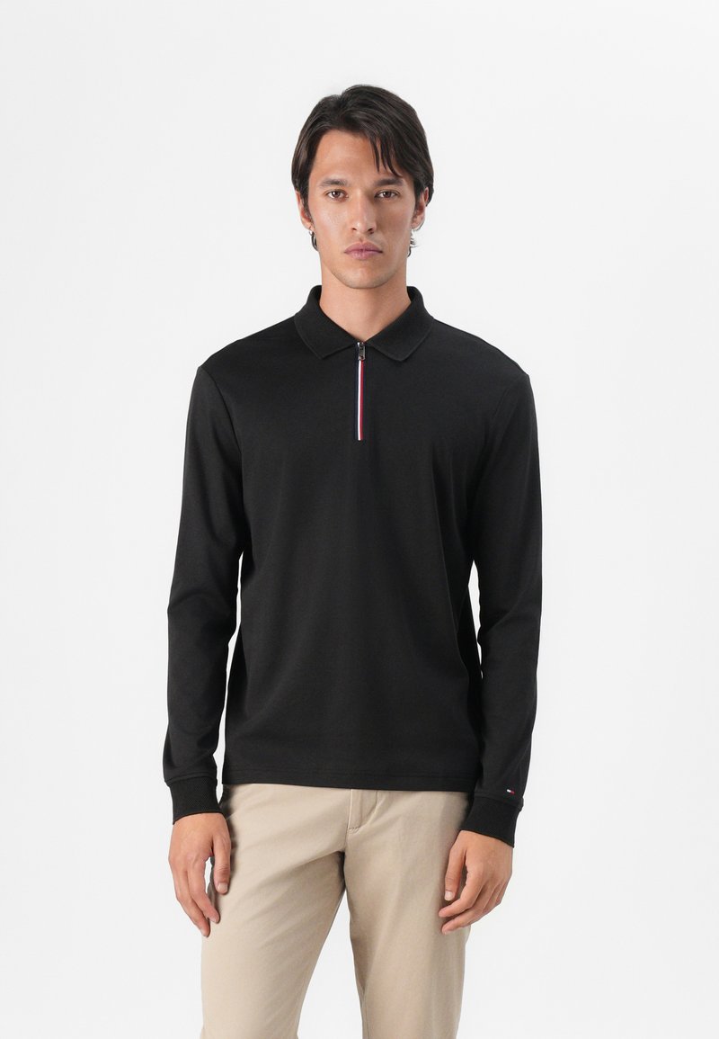 Polo de manga larga negro con cremallera y cuello; presenta un acento de tres rayas en rojo, blanco y azul en la cremallera.