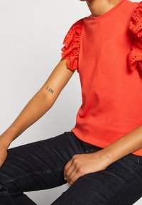 Czerwona koszulka z krótkimi, falbaniastymi, koronkowymi rękawami i okrągłym dekoltem, zestawiona z ciemnymi jeansami. Na przedramieniu widoczny tatuaż.