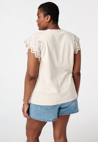 T-shirt en coton beige avec des manches en dentelle à bord festonné. Associé à un short en jean. Texture lisse, design décontracté, montrant la vue arrière.