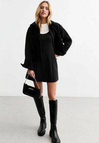 Giacca nera corta con grandi tasche, indossata sopra un vestito nero senza maniche. Stivali neri al ginocchio e una piccola borsa nera con dettagli in catena.
