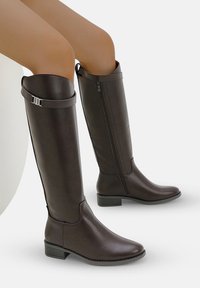 Bottes en cuir marron montantes avec une fermeture éclair sur le côté, accent de boucle réglable et talon bas ; texture lisse et design épuré.