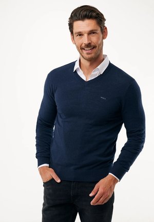 Jersey de cuello en V de color marino hecho de una tela suave y lisa. Presenta puños y dobladillo acanalados, usado sobre una camisa blanca con cuello, con un pequeño logotipo.