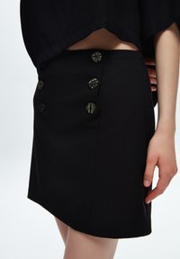 Falda negra con botones dorados estampados, diseño ajustado y textura suave. Cortada por encima de la rodilla, con una abertura lateral para mayor comodidad en el movimiento.