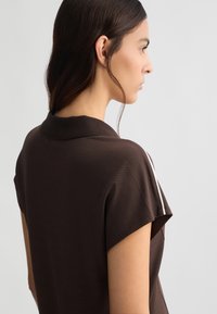 Top corto a maniche corte di colore marrone con colletto, realizzato in un tessuto testurizzato. Una striscia bianca mette in risalto la spalla. Visto di lato.