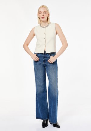 Femme blonde debout portant un haut crème sans manches boutonné avec un col bijou, un jean bleu taille haute à jambes larges et des bottes noires pointues.