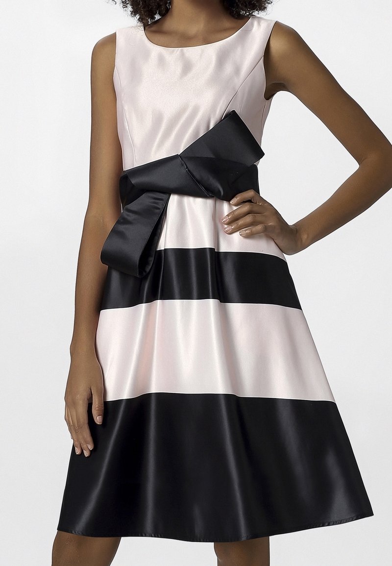 Femme portant une robe sans manches à rayures horizontales rose clair et noir avec un grand nœud noir noué à la taille.