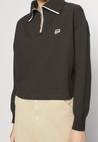 Pullover nero strutturato con zip a quarto, colletto bianco e piccolo logo. Maniche lunghe con polsini, design cropped, abbinato a pantaloni beige.