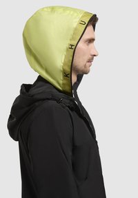 Sort jakke med en lys gul hætte og sort kant. Stoffet ser glat ud, med en lynlås og justerbare snore ved halsen.