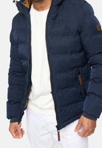 Granatowa kurtka puchowa z zapięciem na zamek, ściągaczami na mankietach i brązowymi skórzanymi akcentami; posiada kaptur i pikowany wzór.