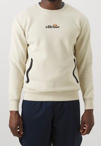 Beige mikina s kulatým výstřihem a žebrovanými manžetami. Obsahuje černé kapsy na zip a vyšívané logo na přední straně.