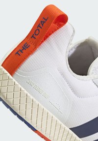 Biele tenisky s textúrovaným zvrškom, s oranžovým pútkom s nápisom "THE TOTAL" v modrej farbe. Výrazná gumová podrážka s vzorovaným gripom.