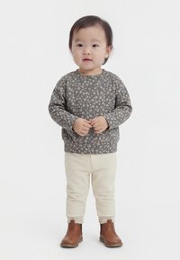 Mazulis stāv, valkājot pelēku ziedu raksta sporta kreklu, krēmkrāsas ribota auduma bikses un brūnus potīšu zābakus uz balta fona.