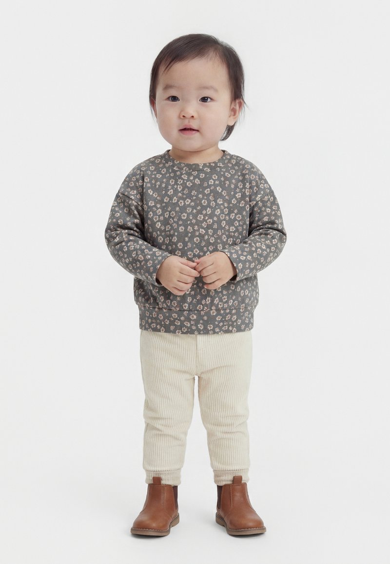 Mazulis stāv, valkājot pelēku ziedu raksta sporta kreklu, krēmkrāsas ribota auduma bikses un brūnus potīšu zābakus uz balta fona.