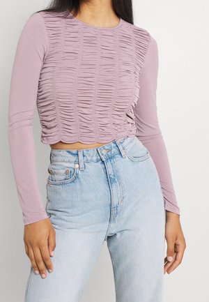 Kobieta ubrana w liliowy, marszczony crop top z długim rękawem, zestawiony z jasnoniebieskimi, wysokim stanem dżinsami na prostym tle.