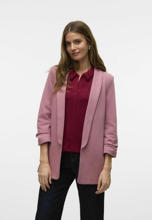 Blazer rose clair au design structuré, avec des manches retroussées et une texture lisse, superposé à un top en tricot torsadé rouge.