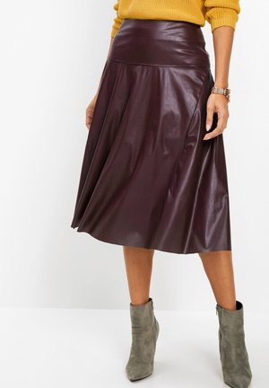 Femme portant une jupe midi en cuir bordeaux, un pull moutarde et des bottines en daim gris, debout devant un fond blanc.