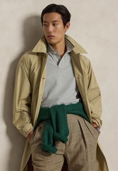 Jeune homme portant un trench-coat beige, un polo gris clair, un pantalon à carreaux et un pull vert noué autour de la taille, debout contre un mur neutre.