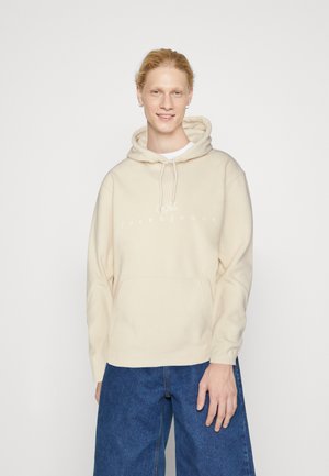Sudadera beige con un bolsillo canguro, capucha ajustable con cordón, y logo blanco. Combinada con pantalones de denim azul de pierna ancha. Textura suave.