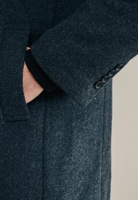 Abrigo de lana gris oscuro con tejido texturizado, que presenta puños de tres botones, bolsillo lateral y un diseño de cuello clásico.
