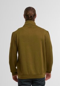 Mann mit zurückgebundenem Haar, der eine olivgrüne, strukturierte Jacke mit geometrischen Mustern trägt, vor einem schlichten hellgrauen Hintergrund steht.