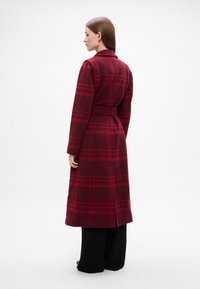 Palton lung cu carouri roșii, prevăzut cu curea, fabricat dintr-un material moale. Are guler și deschideri laterale, purtat peste pantaloni negri largi.