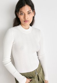 Hvit ribbet langermet topp med høy krage. Stoffet har en glatt og tettsittende tekstur som fremhever vertikale linjer og subtile detaljer.