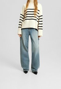 Übergroßer Pullover in Creme und Schwarz gestreift mit hohem Kragen, kombiniert mit weit geschnittenen hellblauen Jeans und spitzen schwarzen Absätzen.