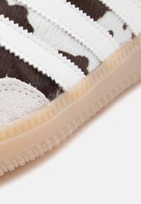 Αθλητικό παπούτσι με λευκό και καφέ σχέδιο αγελάδας, τρεις λευκές ρίγες, καστόρινη μύτη, υφασμένο ελαστικό πέλμα και ραμμένες λεπτομέρειες κατά μήκος των άκρων.