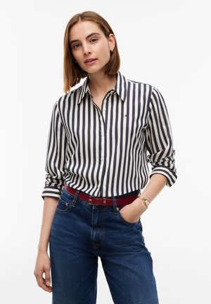 Femme aux cheveux mi-longs portant une chemise à rayures verticales bleu marine et blanche, un jean bleu, une ceinture rouge et une montre en or au poignet gauche.