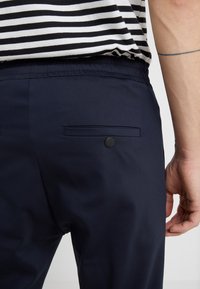 Gestreept zwart-wit shirt ingestopt in marineblauwe broek met een achterzak met een enkele zwarte knop, zichtbare arm met een dunne tatoeage.