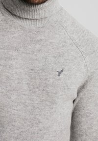 Homme portant un pull à col montant en maille gris clair avec un petit emblème d'oiseau sombre brodé sur la poitrine.