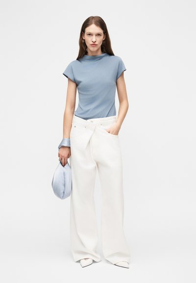 Femme portant un haut à manches courtes bleu clair, un pantalon blanc large, des chaussures blanches pointues, tenant un sac bleu clair avec un accessoire assorti au poignet.