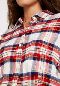 Camicia a quadri flanella rossa, blu e bianca con colletto button-down, caratterizzata da bottoni bianchi e un logo discreto sulla tasca del petto.