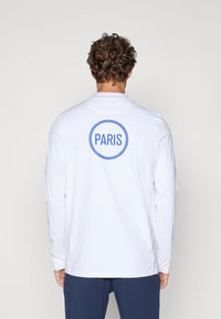 Bijela majica dugih rukava s plavim kružnim logom "PARIS" na leđima, izrađena od laganog materijala s glatkom teksturom.