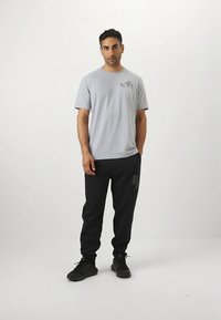 Grijs T-shirt met korte mouwen en een kleine afbeelding op de borst, gecombineerd met zwarte joggingbroeken met een opvallend logo, en zwarte sportschoenen.