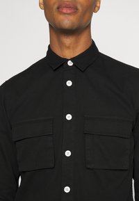 Zwart overhemd met knopen, een puntige kraag, twee borstzakken en witte knopen, met een gladde stoftextuur en een getailleerde pasvorm.