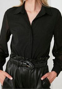 Camicia nera con colletto classico e chiusura a bottoni, abbinata a pantaloni in pelle nera a vita alta e una cintura larga con fibbia rotonda.