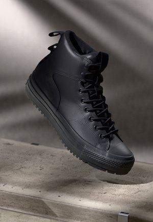 Baskets montantes noires avec une semelle épaisse en caoutchouc et des lacets, flottant au-dessus d'une surface en béton avec des ombres douces en arrière-plan.