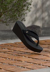 Μαύρη πλατφόρμα σαγιονάρας με πλεκτό λουράκι, που διαθέτει χρυσό έμβλημα. Λαστιχένια σόλα με υφή για καλύτερη πρόσφυση. Τοποθετημένη πάνω σε ξύλινα πλακάκια.