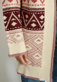 Gebreide cardigan in off-white met rode en roze geometrische patronen. Heeft geribbelde manchetten en zoom, een zachte textuur en franjes aan de randen.